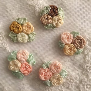 May include: Five handmade fabric flower embellishments in various shades of pink, peach, yellow, and brown. The flowers are arranged in clusters of three and have a delicate, vintage aesthetic.