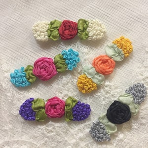 May include: Five handmade fabric flower hair clips in various colors, including red, pink, orange, blue, green, and black. The flowers are made of ribbon and are arranged in a row on a white lace background.