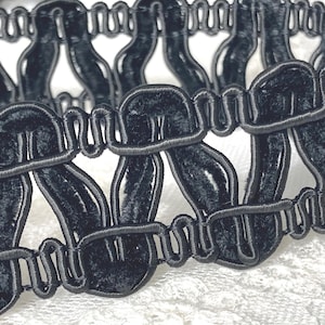 May include: Close-up of a black velvet trim with a looped design. The trim features a series of connected, curved shapes, creating a decorative border. The velvet has a soft texture, and the trim is set against a white background.