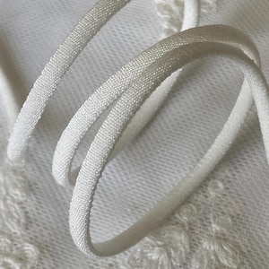 May include: Close-up of white elastic cord, tightly woven with a textured surface. The cord is curved and looped, set against a soft, white, patterned fabric background. The image highlights the cord's texture and color.