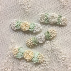May include: Four delicate floral embellishments made of white, cream, and green satin ribbon. The embellishments are arranged in a row on a white lace background.
