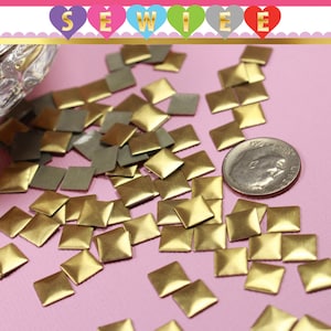 May include: A close-up of a pile of gold and silver square-shaped metal studs. The studs are scattered on a pink surface. A US dime is included for scale.