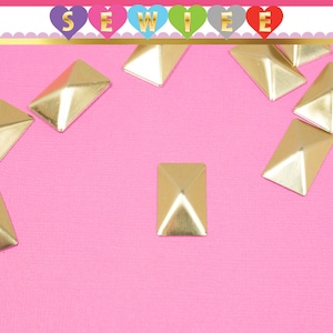 May include: A close-up of gold pyramid-shaped studs on a pink background. The studs are rectangular and have a shiny finish.