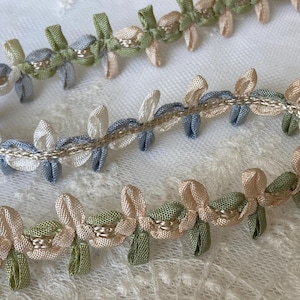May include: Close-up of decorative ribbon trim in pastel colors. The trim features a repeating floral design with light blue, cream, and green ribbon elements, accented with metallic thread. The ribbon is arranged in a wavy pattern.