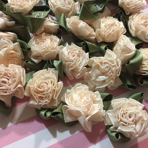 May include: A collection of small, peach-colored fabric flowers with green ribbon bows. The flowers are arranged in a cluster on a pink and white striped surface.