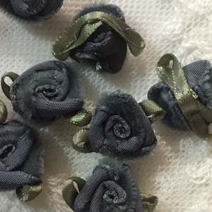 May include: A collection of gray fabric rose embellishments with green ribbon accents. The roses are sewn together in a spiral shape and have a soft, velvety texture.