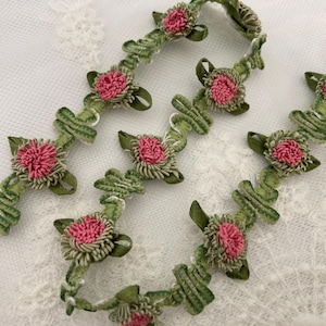 May include: Close-up of decorative floral trim. The trim features pink, textured flower centers surrounded by green leaves and stems. The trim is arranged on a white, lace-like fabric.