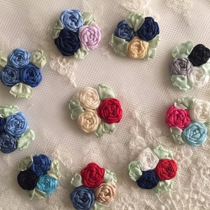 May include: Assortment of 10 handmade fabric flower embellishments in various colors, including blue, red, pink, white, and cream. Each flower is made of ribbon and has a green leaf accent.