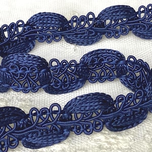 May include: Close-up of navy blue decorative trim. The trim features a series of oval-shaped woven elements connected by intricate, looped designs. The trim is made of a soft, possibly fabric material, and is set against a white background.