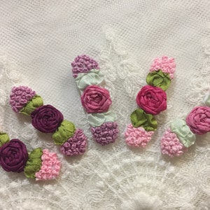 May include: Five handmade fabric flower embellishments in shades of pink, purple, and green. The flowers are made of ribbon and have a 3D effect.