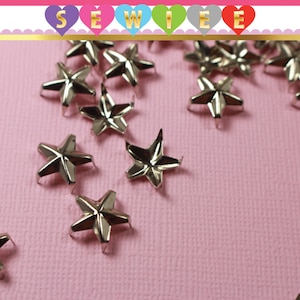 May include: A close-up of silver star-shaped metal studs on a pink surface. The studs have a pointed base and are designed for attaching to fabric.