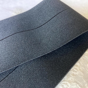 May include: Close-up of a folded, dark gray elastic band. The band has a woven texture and a subtle sheen. The edges are neatly finished, and the band appears to be durable. The background is a white, textured fabric.
