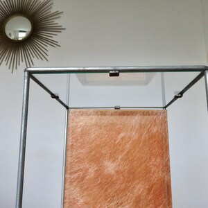 May include: A display cabinet with a clear glass top and sides, supported by a silver metal frame. The interior features a textured, orange-brown panel. A gold sunburst mirror and a small, round, green wall decoration are visible in the background.