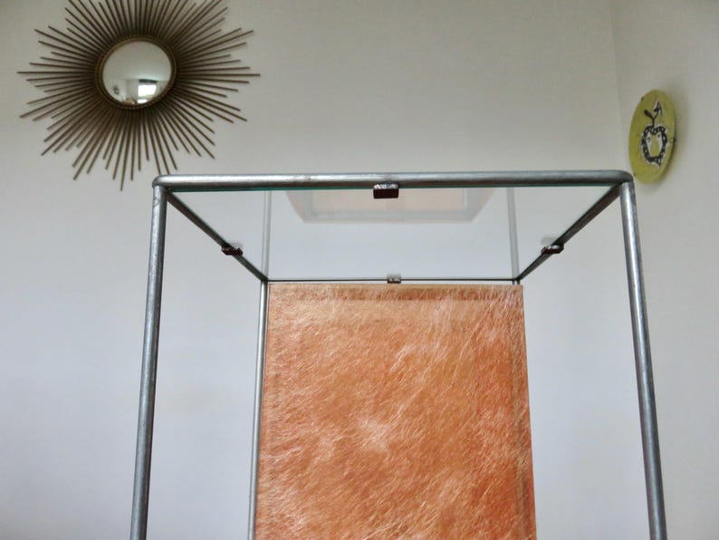 May include: A display cabinet with a clear glass top and sides, supported by a silver metal frame. The interior features a textured, orange-brown panel. A gold sunburst mirror and a small, round, green wall decoration are visible in the background.