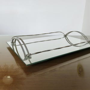 Puede incluir: Una bandeja rectangular con espejo y asa de metal plateado. La bandeja tiene una superficie reflectante y está diseñada con una estética moderna. Las asas son curvas y añaden un elemento decorativo a la bandeja.