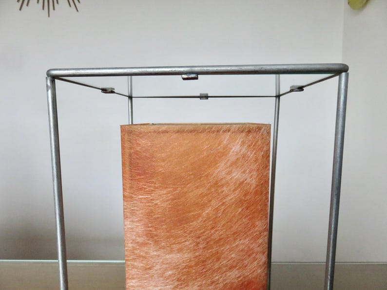 May include: A rectangular, brown, textured object is encased within a silver metal frame. The object has a light brown, fur-like appearance. The frame is a simple, geometric design, with vertical and horizontal bars.