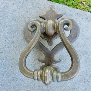 May include: Bronze-colored door knocker with a decorative design. The knocker features a curved handle and ornate backplate with a starburst and heart-shaped elements. The hardware is mounted on a gray surface.