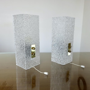 May include: A pair of rectangular table lamps featuring clear, textured glass bodies with a rough, ice-like finish. Each lamp includes a vertical brass-toned metal plate mounted on the front with two small spherical knobs and a white pull cord.