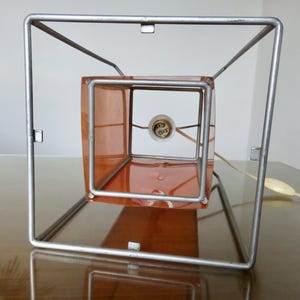 May include: A contemporary table lamp featuring a square metal frame. The lamp has a translucent orange cube inside, housing the light bulb. The design is simple, with a power cord and switch. The lamp sits on a wooden surface.