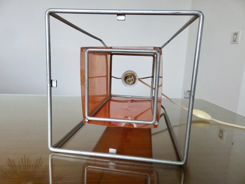 May include: A contemporary table lamp featuring a square metal frame. The lamp has a translucent orange cube inside, housing the light bulb. The design is simple, with a power cord and switch. The lamp sits on a wooden surface.