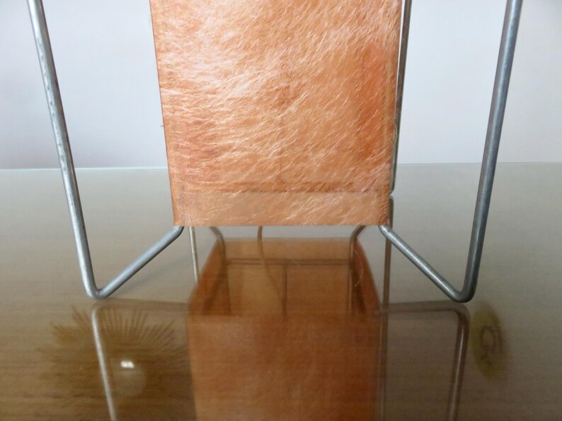May include: A retro table lamp featuring a rectangular shade and a metal frame. The shade is a warm orange hue with a textured, fibrous appearance. The metal frame is silver and has a minimalist design.