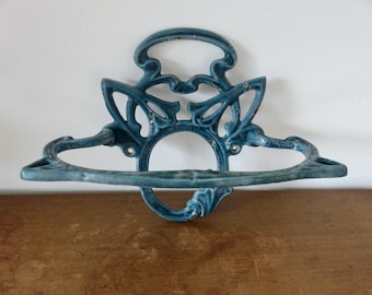 Puede incluir: Un toallero de pared de metal color verde azulado con un marco simétrico de curvas complejas que presenta signos visibles de desgaste y pintura descascarada. Incluye un anillo circular central y una placa decorativa.