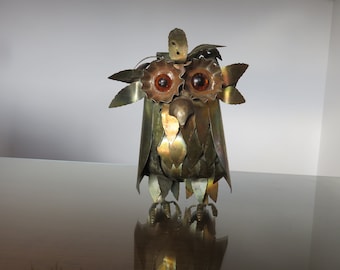 May include: A metal owl sculpture with a brown and gold finish. The owl has large, round, amber-colored eyes and is made of many small, overlapping pieces of metal.