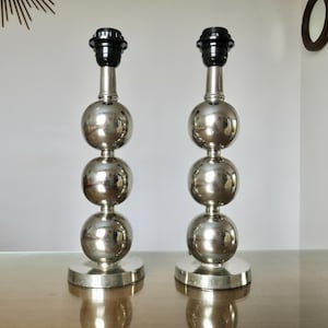 May include: A pair of silver-colored metal table lamp bases, each featuring a vertical stack of three polished spherical orbs mounted on a circular base, topped with a black plastic light bulb socket fixture for standard lamp shades.