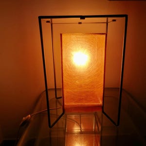 May include: A rectangular table lamp with a warm orange glow. The lamp has a black metal frame and a textured orange shade. The light source is visible through the shade, creating a cosy ambiance. The lamp is on a glass surface.