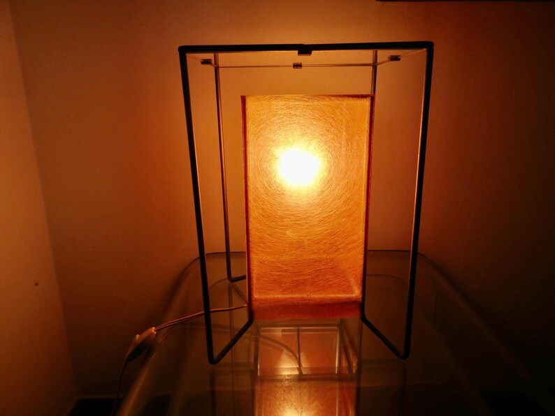 May include: A rectangular table lamp with a warm orange glow. The lamp has a black metal frame and a textured orange shade. The light source is visible through the shade, creating a cosy ambiance. The lamp is on a glass surface.