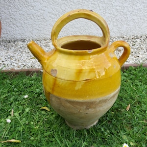 May include: A yellow and tan ceramic pitcher with a handle and spout. The pitcher has a rounded body and a wide opening. It is sitting on green grass.