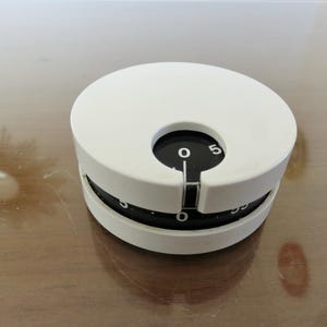 Peut inclure: Un minuteur de cuisine blanc avec un cadran noir et un design circulaire. Le minuteur a un indicateur noir et affiche des chiffres de 0 à 5. Le minuteur est sur une surface en bois.