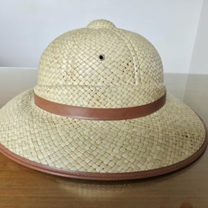Peut inclure: Un casque colonial en paille tressée beige clair, doté d'une calotte arrondie avec un petit œillet de ventilation et d'un large bord incliné vers le bas. Le chapeau est orné d'une bande marron lisse entourant la base de la calotte.