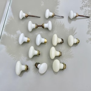 May include: A collection of twelve white porcelain door knobs with brass-toned metal hardware. The set includes various individual knobs and one double-sided knob assembly, all featuring smooth, rounded surfaces and metallic mounting components.