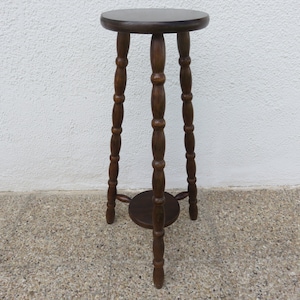 Può includere: Un portapiante in legno marrone scuro con piano rotondo e tre gambe tornite. Il supporto ha un piccolo ripiano rotondo vicino alla base delle gambe. Il supporto è alto circa 71 cm.