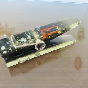 May include: A large clothespin featuring a translucent black and yellow gradient resin body. Embedded within the resin are small white seashells and a prominent orange seahorse figure. A silver-toned metal spring mechanism connects the two halves.