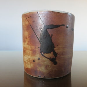 Puede incluir: Una taza de cerámica marrón con un diseño abstracto negro. La taza tiene un esmalte moteado y una forma ligeramente redondeada.