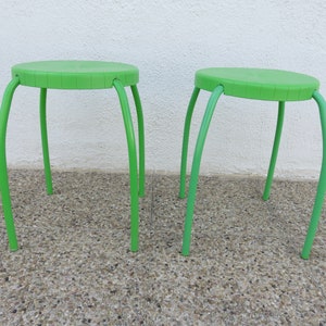 May include: Two green metal stools with round seats and three legs each. The stools are on a gray concrete surface.