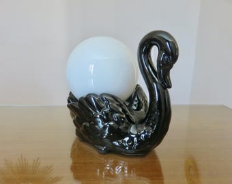 May include: A black ceramic swan ornament supporting a white globe light. The swan has a shiny finish and sits on a wooden surface. The light is a bright, opaque white.