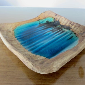 May include: A square, handmade ceramic dish with a textured, light brown exterior and a vibrant blue interior. The interior features a glossy, rippled design, creating a water-like effect. The dish is ideal for serving or display.