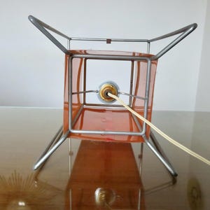 May include: A vintage table lamp featuring an orange square shade within a silver metal frame. The lamp has a four-legged base and a power cord. The lamp is on a reflective surface.