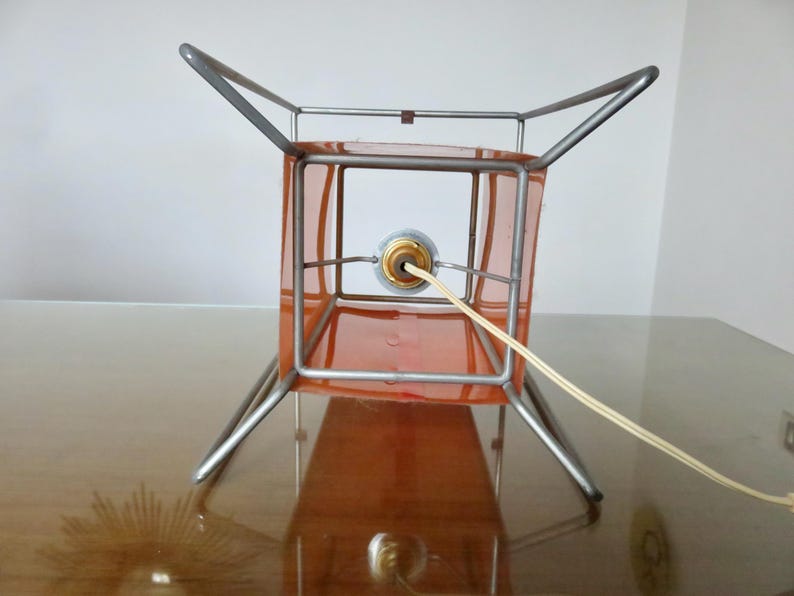 May include: A vintage table lamp featuring an orange square shade within a silver metal frame. The lamp has a four-legged base and a power cord. The lamp is on a reflective surface.