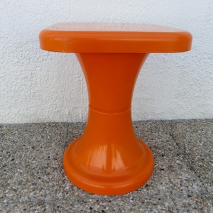 Op de afbeelding: Een feloranje kruk met een gladde, glanzende afwerking. De kruk heeft een vierkant blad met afgeronde hoeken en een centrale voet die naar onderen toe uitloopt, wat zorgt voor een opvallend zandloperachtig silhouet in een effen kleur.
