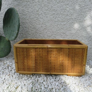 Puede incluir: Un macetero rectangular de mimbre tejido de color marrón natural. El macetero está hecho de bambú tejido y tiene un acabado liso y pulido.