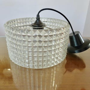 Könnte beinhalten: Transparente Glas-Pendelleuchte mit geometrischem Muster. Die Leuchte hat ein schwarzes Kabel und eine schwarze Deckenhalterung. Der Schirm ist zylindrisch und hat eine strukturierte, facettierte Oberfläche. Für Deckenbeleuchtung.