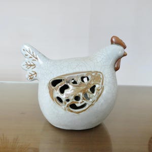 Op de afbeelding: Een keramieken kippenbeeldje met een witte craquelé glazuurafwerking. Het stuk heeft een bruine kam en lellen, met een decoratief opengewerkt vleugelontwerp in een metallic bronskleur. De staartveren zijn versierd met bruine bladpatronen.