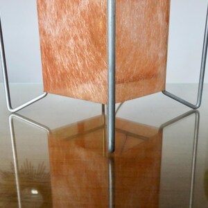 May include: A contemporary table lamp featuring a rectangular shade and a silver metal frame. The shade has a textured, orange-brown surface. The lamp is reflected in the glossy surface below.