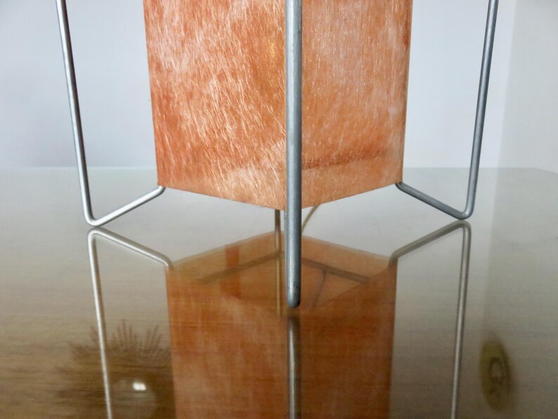May include: A contemporary table lamp featuring a rectangular shade and a silver metal frame. The shade has a textured, orange-brown surface. The lamp is reflected in the glossy surface below.