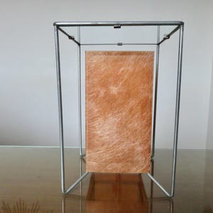 May include: A contemporary lamp with a metal frame. The lamp has a rectangular, orange-brown panel. The lamp is designed to sit on a table or other surface. The frame is made of brushed metal.