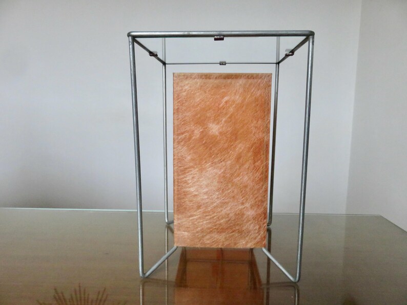 May include: A contemporary lamp with a metal frame. The lamp has a rectangular, orange-brown panel. The lamp is designed to sit on a table or other surface. The frame is made of brushed metal.